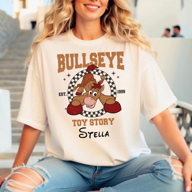 Może przedstawiać: Biały t-shirt z kr&oacute;tkim rękawem z grafiką przedstawiającą postać Bullseye z Toy Story. Wz&oacute;r zawiera szachownicowe koło, napis BULLSEYE, EST. 1999, TOY STORY oraz imię Stella zapisane czarną kursywą pod gł&oacute;wną grafiką.