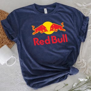 May include: Navy blue t-shirt featuring the Red Bull logo. The logo includes two red bulls facing each other in front of a yellow circle, with the words "Red Bull" in red below. The shirt is made of a soft material.