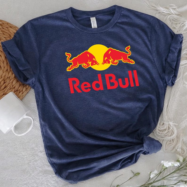 May include: Navy blue t-shirt featuring the Red Bull logo. The logo includes two red bulls facing each other in front of a yellow circle, with the words "Red Bull" in red below. The shirt is made of a soft material.