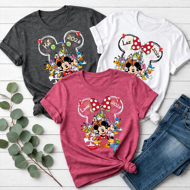 May include: Three short-sleeved t-shirts in gray, white, and pink, each featuring a Disney-themed graphic. The design includes Mickey Mouse, Minnie Mouse, and other characters, with the text "2026" and names. A pair of blue jeans is also visible.
