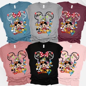 May include: Assorted-color Disney-themed t-shirts. Each shirt features a Mickey Mouse head outline with a name and the year 2026. The shirts display Mickey Mouse, Minnie Mouse, and other Disney characters. The shirts are in various colors.