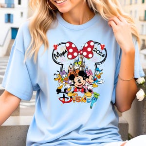 May include: Light blue t-shirt featuring a colorful Disney graphic. The design includes Mickey and Minnie Mouse, Donald Duck, Goofy, and other characters. The word "Disney" is displayed in a stylized font, with the text "Mapa 2026" above the characters.