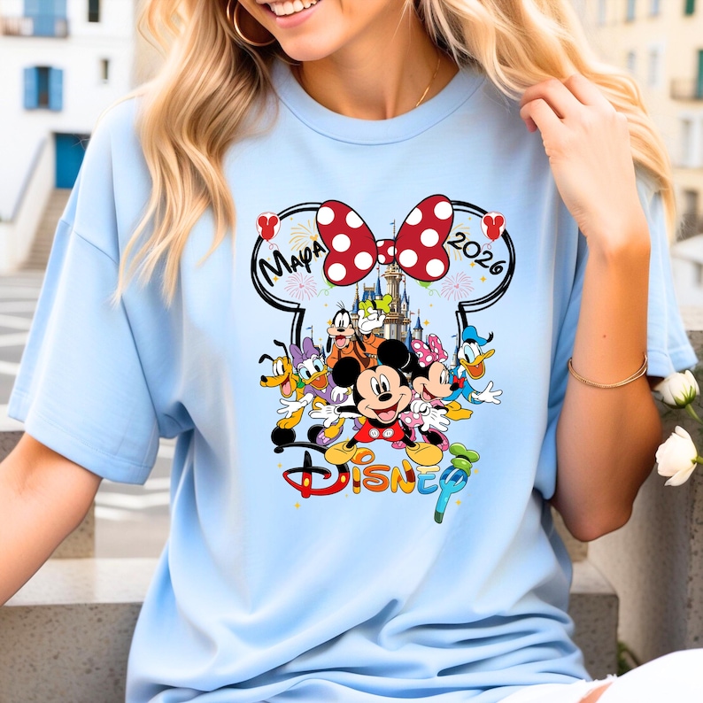 May include: Light blue t-shirt featuring a colorful Disney graphic. The design includes Mickey and Minnie Mouse, Donald Duck, Goofy, and other characters. The word "Disney" is displayed in a stylized font, with the text "Mapa 2026" above the characters.