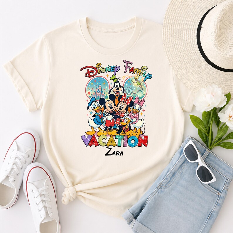 May include: Cream-colored t-shirt featuring a colorful Disney Family Vacation graphic. The design includes Mickey Mouse, Minnie Mouse, Donald Duck, Daisy Duck, Goofy, and Pluto. The word "VACATION" is prominently displayed.