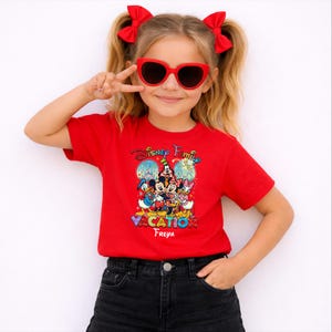 May include: A red t-shirt featuring a colorful graphic of Disney characters, including Mickey and Minnie Mouse, Donald Duck, and Daisy Duck. The shirt has the text "Disney Family Vacation" and the name "Freya" below the graphic. The person is wearing red heart-shaped sunglasses.