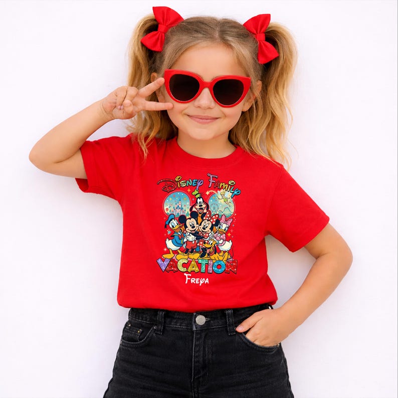 May include: A red t-shirt featuring a colorful graphic of Disney characters, including Mickey and Minnie Mouse, Donald Duck, and Daisy Duck. The shirt has the text "Disney Family Vacation" and the name "Freya" below the graphic. The person is wearing red heart-shaped sunglasses.