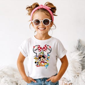 May include: A white t-shirt featuring a colorful Disney graphic with Mickey Mouse, Minnie Mouse, and other characters. The design includes the text "Vera 2026" and the Disney logo. The shirt is worn by a young girl.
