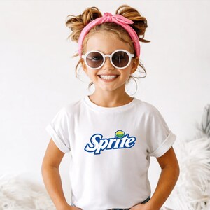 May include: A young person wearing a white t-shirt with the "Sprite" logo in blue and green. The person is also wearing white sunglasses and a pink headband. The image is well-lit and the person is smiling.