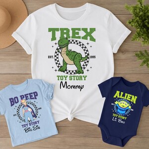 Może przedstawiać: Biały t-shirt z grafiką T-Rexa i napisem TREX TOY STORY Mommy EST 1995, jasnoniebieski t-shirt z Bo Peep i napisem BO PEEP TOY STORY Big Sis EST 1995 oraz granatowe body z kosmitą i napisem ALIEN TOY STORY Lil Bro EST 1995.