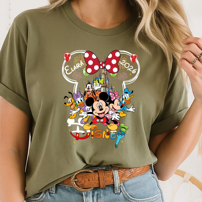 May include: Olive green t-shirt featuring a colorful Disney graphic. The design includes Mickey Mouse, Minnie Mouse, Donald Duck, and other characters. The word "Disney" is displayed in a playful font, with the name "Elara" and the year "2026" above the characters.