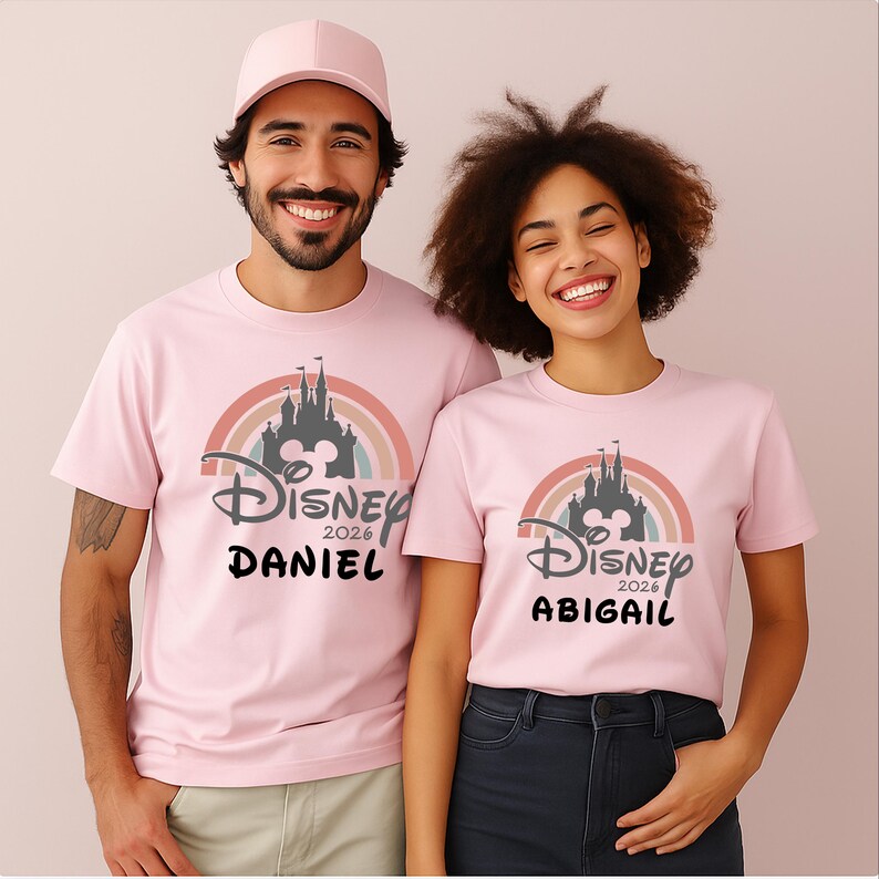 May include: Pink t-shirts with a Disney castle design, rainbow, and "Disney 2026" text. One shirt says "Daniel", the other "Abigail". The shirts are worn by two people.