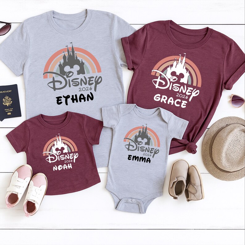 May include: Four Disney-themed tops and a babygrow in grey and burgundy. Each item displays a castle design with a rainbow and the text "Disney 2026" plus a name. Accessories include sunglasses, a hat, and shoes.