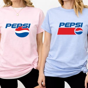 May include: Two short-sleeved t-shirts, one light pink and one light blue, each featuring the Pepsi logo. The logo includes the word "PEPSI" in blue, with a red and blue design. The shirts are a casual style.