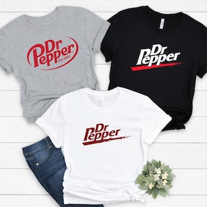 May include: Three t-shirts with the Dr Pepper logo in red and white. One shirt is gray with the logo in a circular design and the text "Est. 1885". Another shirt is black with the logo and a red stripe. The third shirt is white with the logo and a red stripe.