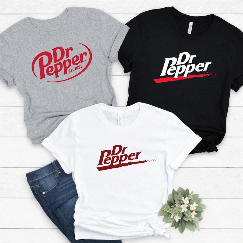 May include: Three t-shirts with the Dr Pepper logo in red and white. One shirt is gray with the logo in a circular design and the text "Est. 1885". Another shirt is black with the logo and a red stripe. The third shirt is white with the logo and a red stripe.