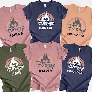 May include: Six short-sleeved t-shirts in different colours, each with a Disney castle design, a rainbow, and the text "Disney 2026" and a name. The colours are pink, navy, orange, green, and blue.