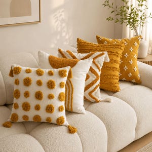 May include: A selection of decorative cushions in mustard yellow and white. The cushions feature various patterns, including dots, chevrons, and floral designs. Some cushions have tassels. The cushions are arranged on a white sofa, creating a cozy aesthetic.