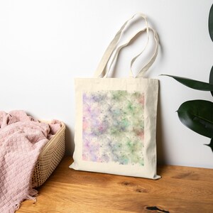 May include: A cream-coloured canvas tote bag featuring a square central print with a soft, abstract watercolour pattern in shades of pastel purple, green, and pink, complete with two long fabric handles for carrying over the shoulder.
