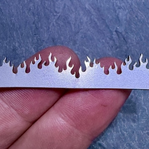 May include: A close-up of a silver metal strip with a flame-like design cut along the top edge. The strip is held between fingers, with a blue-grey background. The flames are evenly spaced, creating a decorative border.