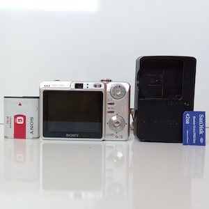 Può includere: Una fotocamera digitale Sony DSC-W50 color argento con schermo LCD posteriore, ghiera delle modalità e pulsanti di menu. Sono inclusi una batteria agli ioni di litio Sony NP-BG1 bianca, un caricabatterie nero e una scheda SanDisk Memory Stick PRO Duo blu da 2 GB.
