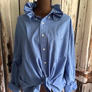 Peut inclure: Chemise boutonnée bleu clair à carreaux avec col à volants, nouée à la taille. Les manches sont retroussées et la chemise est présentée sur un mannequin. Boutons blancs.