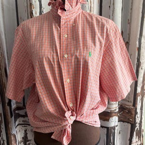 Puede incluir: Una camisa de manga corta con botones, un patrón de cuadros naranja y blanco, un cuello alto con volantes y un pequeño logotipo bordado en verde en el pecho izquierdo. La camisa está anudada en la cintura y tiene botones claros al frente.