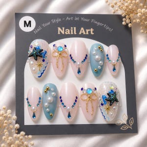 May include: A set of oval-shaped nail art in various designs. The nails feature light pink and blue bases, adorned with pearls, rhinestones, and gold accents. Some nails have a starfish design. The package reads "Nail Art".