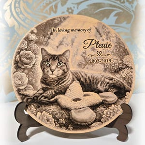 May include: A circular wooden plaque featuring a laser-engraved portrait of a tabby cat lying next to a toy. The text reads In loving memory of Ptuie 2003-2019. The design includes floral elements and is displayed on a dark brown wooden stand.