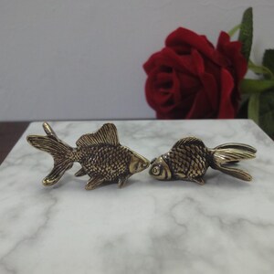 May include: Two small, metallic, brass-colored fish figurines with detailed scale textures and fins. The fish are positioned facing each other, showcasing a weathered, antique-style finish on their surfaces. These decorative items are solid.