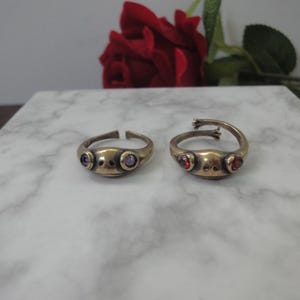 May include: Two open-band rings crafted from a metallic material, each shaped like a frog head. The left ring features two small purple circular stones as eyes, while the right ring displays two red circular stones as eyes with visible frog feet.