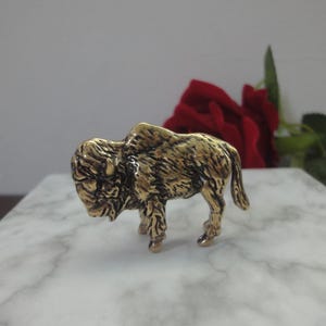 May include: A small, metallic gold-toned figurine of a bison standing in profile. The sculpture features detailed, textured fur patterns across its body, a prominent hump on its back, and a downward-facing head, capturing the animal's sturdy form.