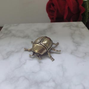 May include: A metallic gold-toned beetle figurine with a smooth, rounded shell and six articulated legs. The insect sculpture features a segmented body design with a polished finish, capturing the anatomical form of a scarab beetle in a stationary pose.