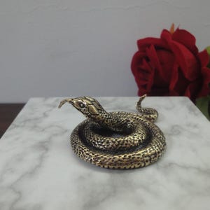 Puede incluir: Una pequeña figura de serpiente enroscada hecha de un material metálico con un acabado en tono latón. La superficie presenta un patrón texturizado similar a escamas en todo el cuerpo, con la cabeza levantada y la lengua extendida hacia adelante.