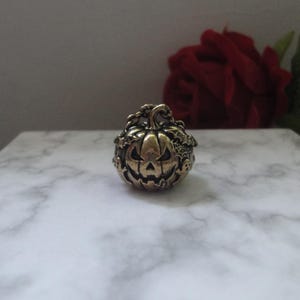 May include: A metallic, brass-toned ring featuring a sculpted jack-o'-lantern pumpkin design. The pumpkin has a carved, grinning face with triangular eyes and a jagged mouth, surrounded by small, raised details of bats and spider webs.