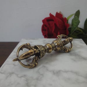 May include: A metallic gold-toned vajra ritual object featuring intricate symmetrical carvings, a central spherical grip, and multi-pronged ends that curve inward to meet at a pointed tip, showcasing a polished finish with detailed metalwork.