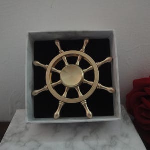 Op de afbeelding: Een goudkleurige metalen fidget spinner in de vorm van een scheepsroer met acht spaken en een glad rond midden. Het object wordt gepresenteerd in een vierkante witte doos met een zwarte fluwelen inzet, wat een duidelijk zicht op het ontwerp geeft.
