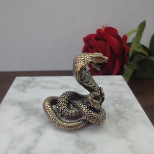 May include: A small, metallic, brass-coloured cobra figurine in a coiled, upright striking position. The snake features detailed, textured scales across its body and a flared hood. The sculpture is rendered in a weathered, antique-style finish.