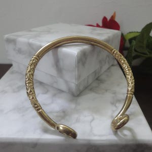 May include: A gold-toned metal cuff bracelet featuring a smooth, rounded band with two distinct sections of intricate, embossed scrollwork patterns. The bracelet has an open design with rounded, polished ends that curve slightly inward.