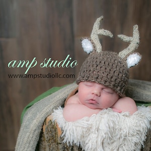 May include: A newborn baby wearing a brown knitted deer hat with white antlers and white fur trim, sleeping in a wooden log with a green and brown blanket.