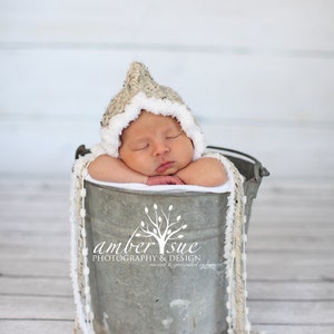May include: A newborn baby wearing a crocheted hat with a white trim is sleeping in a metal bucket. The bucket is decorated with white and beige yarn. The background is a white wood panel.