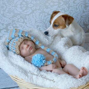 May include: A newborn baby wearing a brown and blue crocheted hat with a blue pom-pom lies in a wicker basket with a white blanket. A small white and brown dog looks at the baby.