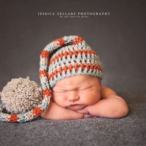 May include: A newborn baby wearing a handmade crochet hat with a long, pointed top and a large pom-pom. The hat features stripes of orange, light blue, and cream colors. The baby is lying on a gray surface.