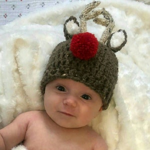May include: A baby wearing a brown crocheted reindeer hat with white antlers, a red pom-pom, and white felt ears.
