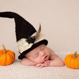 May include: A newborn baby wearing a black witch hat with a white burlap bow and a feather accent, sleeping on a beige textured surface with two orange pumpkins.