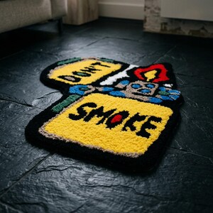 May include: A black, yellow, and red rug shaped like a lighter. The rug has the words "DON'T" and "SMOKE" in black lettering on yellow rectangles. The flame is red and yellow, with blue and green accents.