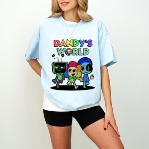 May include: Pale blue t-shirt with the text "Dandy's World" in rainbow colours and a cartoon graphic of three characters. The characters include a TV-headed figure, a flower-like character, and a blue-hatted character holding a small pot.
