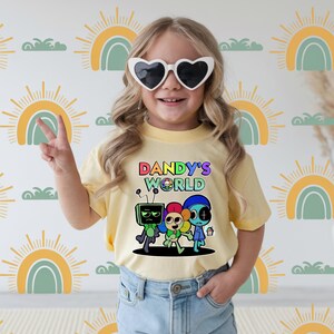 May include: A pale yellow t-shirt featuring the words "DANDY'S WORLD" and a vibrant cartoon illustration. The graphic shows three characters in various colours. The person is wearing heart-shaped sunglasses and blue denim jeans.