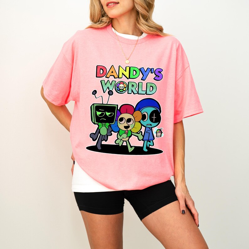 May include: A light pink t-shirt featuring the text "DANDY'S WORLD" in rainbow hues and a cartoon graphic of three characters. The characters include a green TV head, a flower-like character, and a blue character holding a small cupcake.