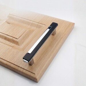 Peut inclure: Une poign&eacute;e de meuble moderne avec un centre noir et un accent argent&eacute; poli. La poign&eacute;e est mont&eacute;e sur une porte d'armoire en bois de couleur claire. La poign&eacute;e mesure environ 15 cm de long.