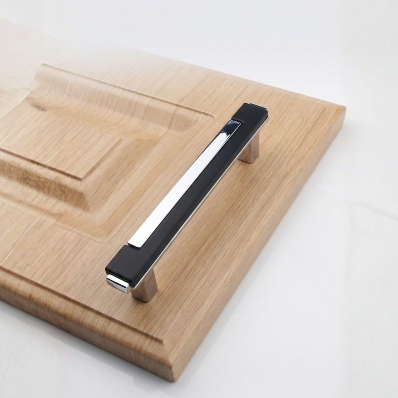 Peut inclure: Une poign&eacute;e de meuble moderne avec un centre noir et un accent argent&eacute; poli. La poign&eacute;e est mont&eacute;e sur une porte d'armoire en bois de couleur claire. La poign&eacute;e mesure environ 15 cm de long.